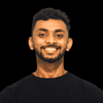 A young man with short curly hair and a beard smiles at the camera. He is wearing a plain black shirt, posed against a solid black background—an ideal look for modern corporate video production.